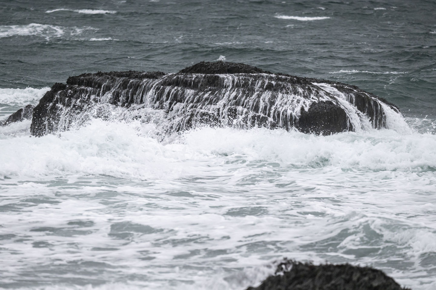 A large rock in the water has many rivulets falling off of it, the aftermath of a wave.