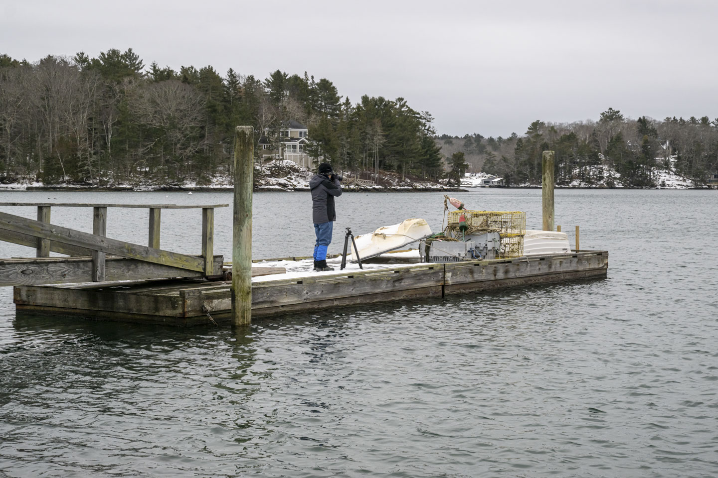 Anne is standing on the dock, wearing a winter coat and holding a camera to her eye.