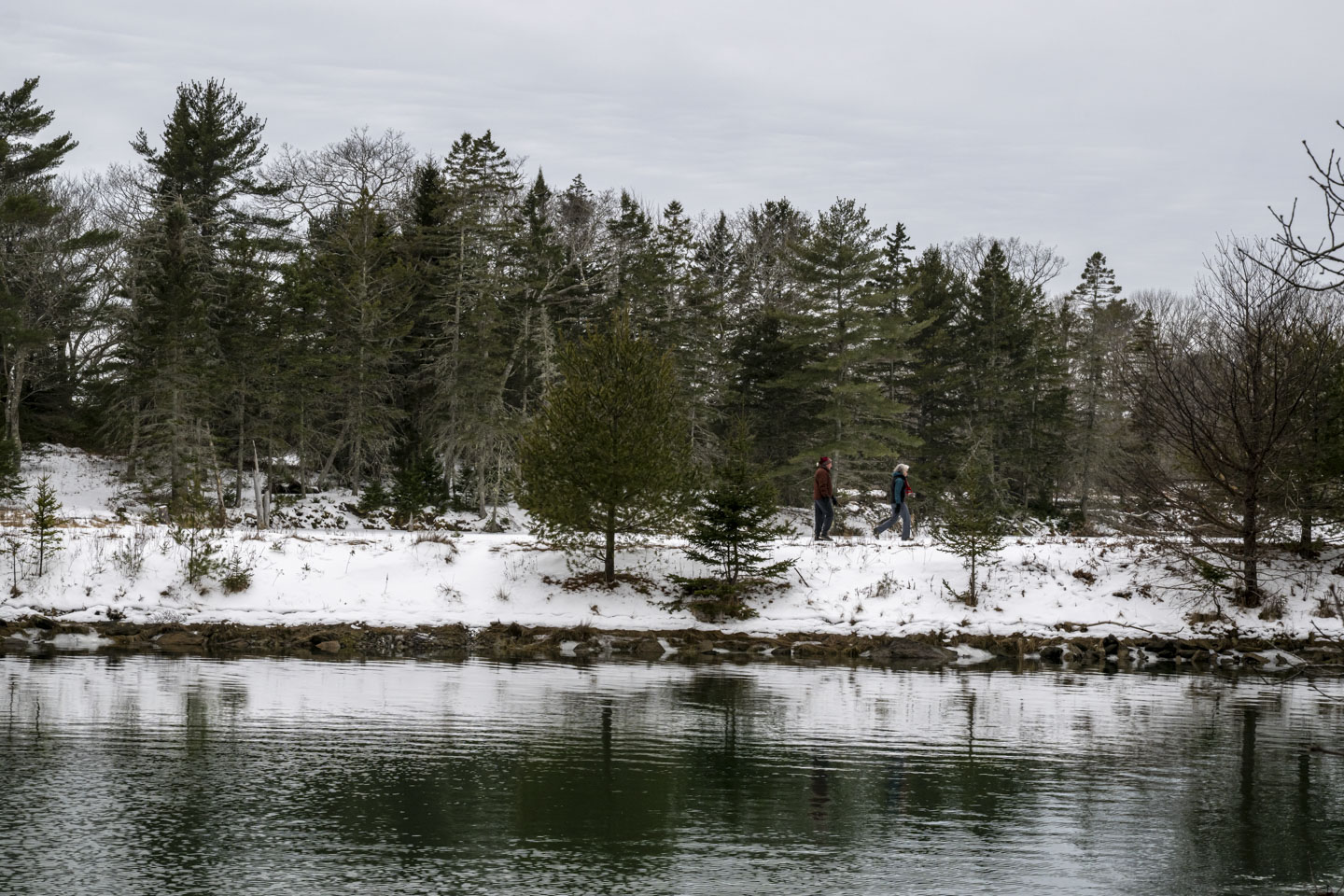 Looking across water, a snow-covered embankment has two people walking on it.