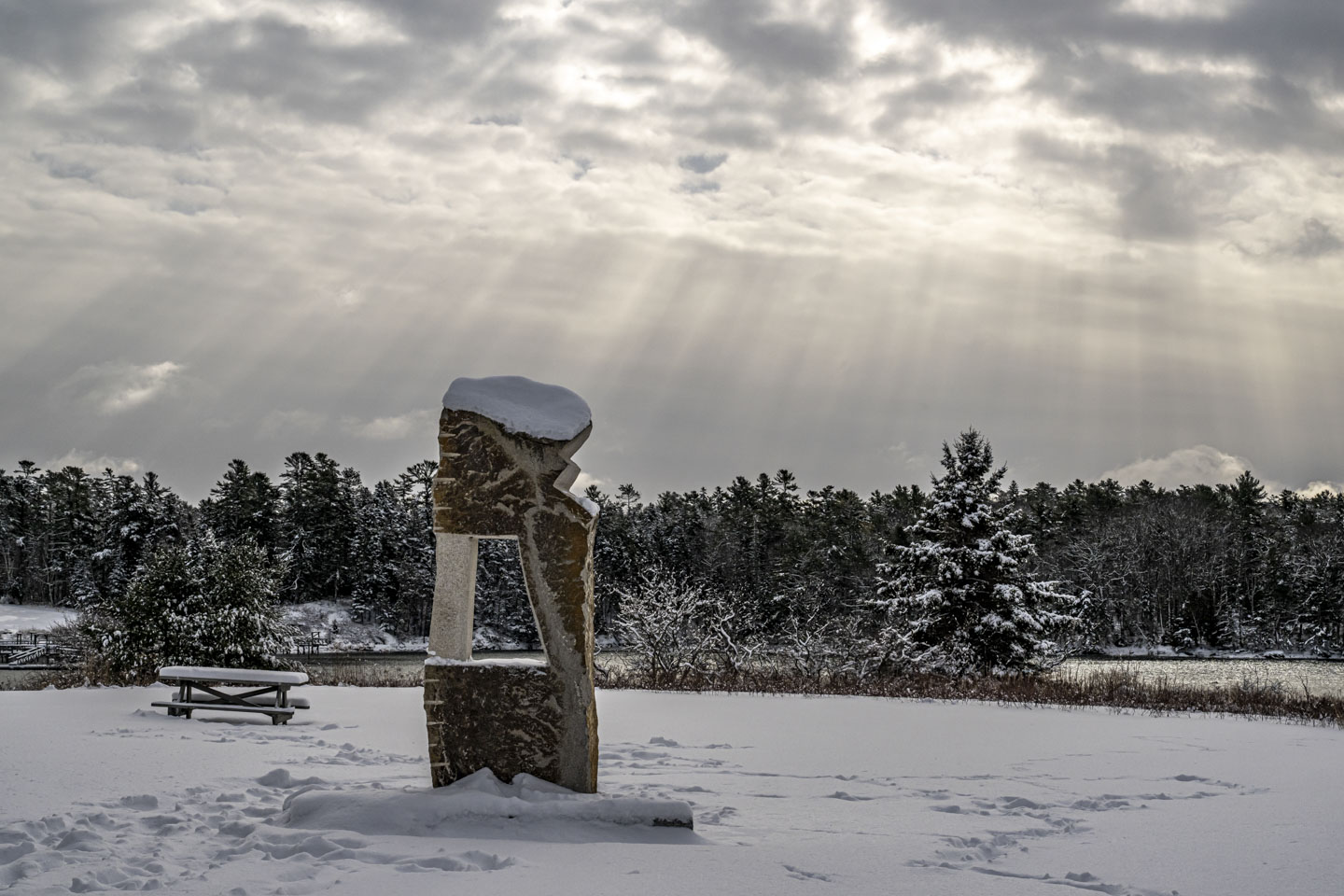 Sunlight streams from a cloud-covered sky on to a snow-covered field that has an abstract blocky sculpture in the middle of it.