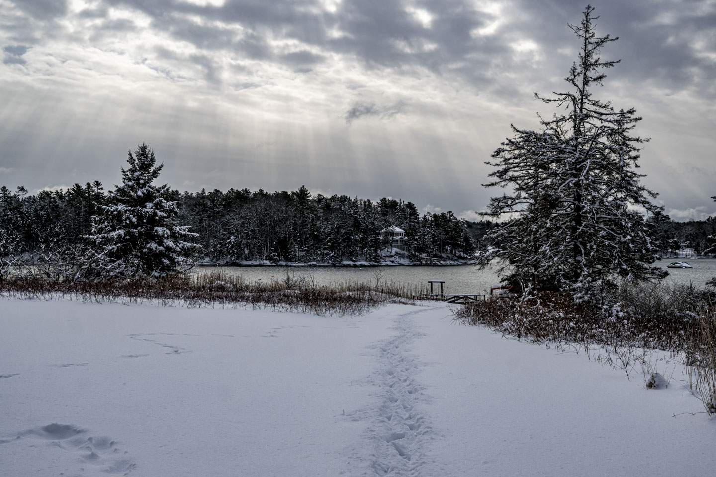 A path through the snow leads to a pier and water, while above sunlight streams through openings in the clouds.