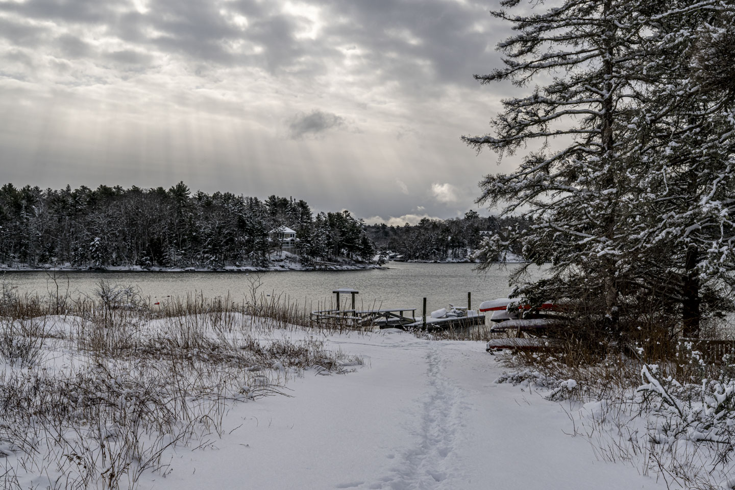 A path through the snow leads to a pier and water, while above sunlight streams through openings in the clouds.