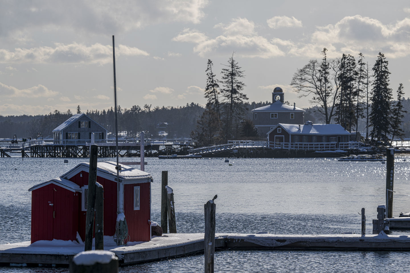 A red shed is on a floating dock. Everything has a bit of snow on it. A house on stilts can be seen in the background, as well as a couple houses on an island.