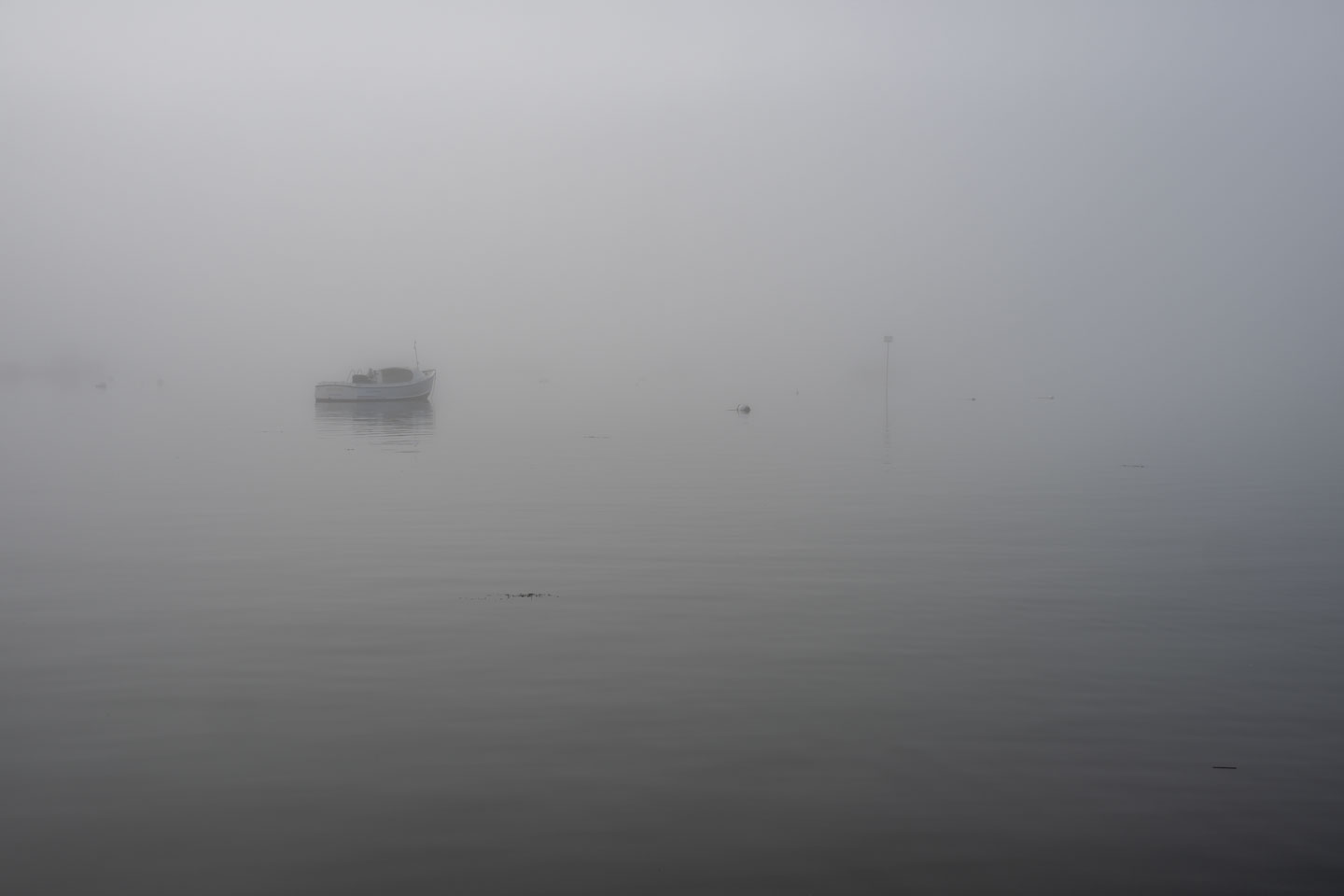 A dense fog almost obscures a boat that is on the water, with early morning light diffused by the fog.