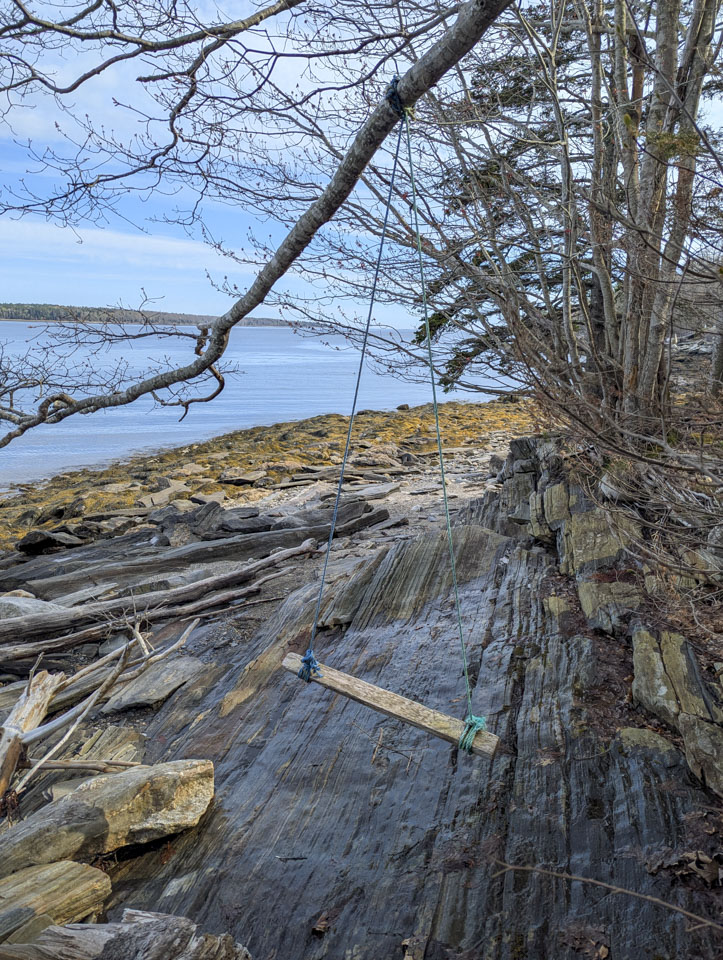A thin board is hanging via twine from a rather thin tree branch. Wet rock is underneath and a river is in the background.