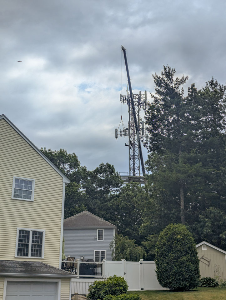 Worker leaning from a cell tower while a crane lifts equipment above houses and trees under a cloudy sky