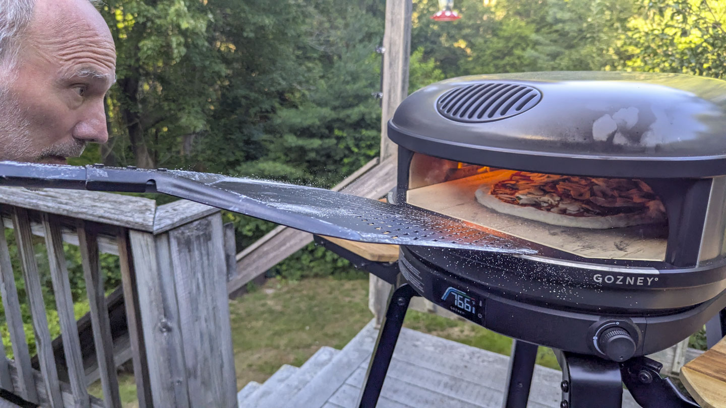 Paul is peering into the oven, with a black pizza peel being pulled from it.