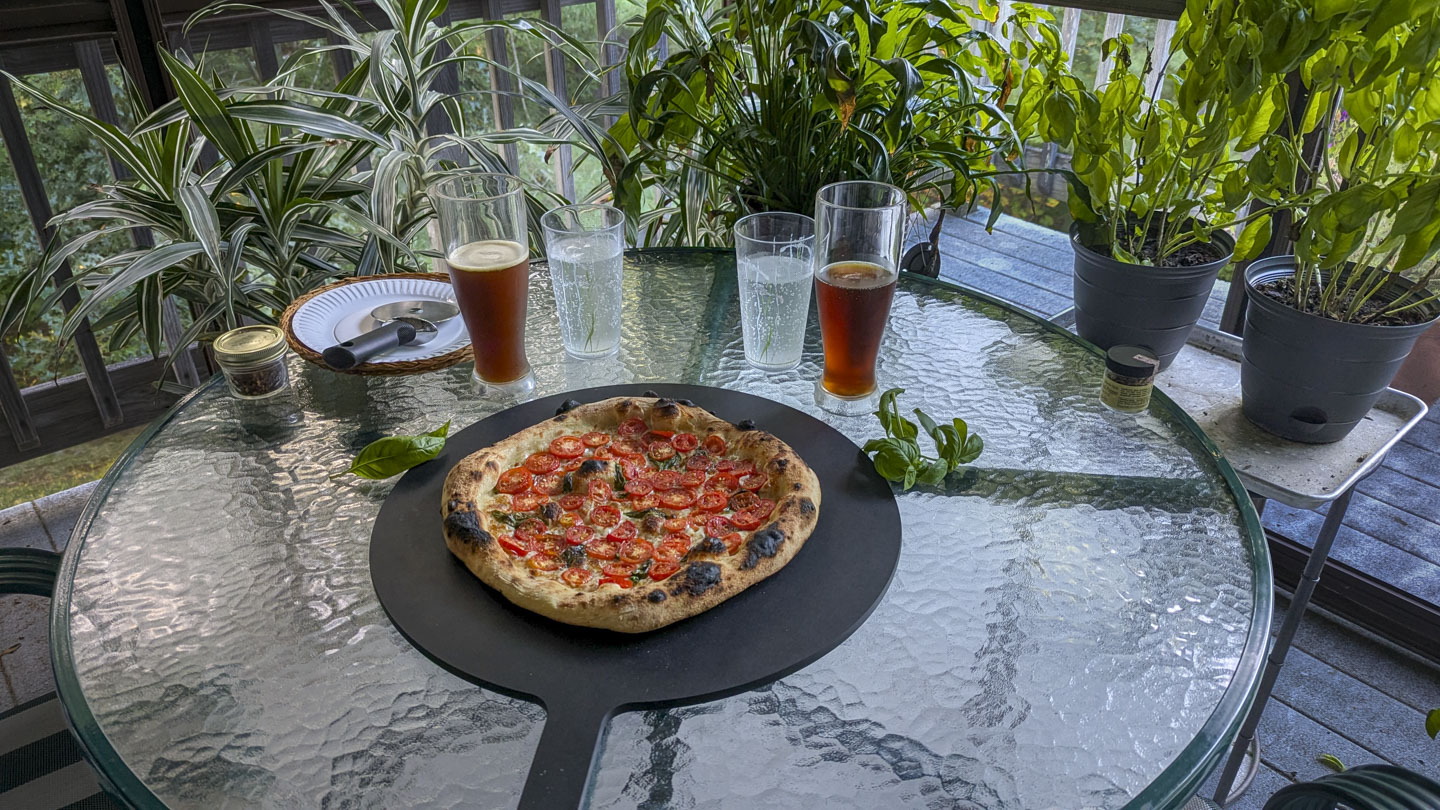 A nicely charred pizza with sliced tomatoes on it sits on a black cutting board on a glass table with plants in the background.