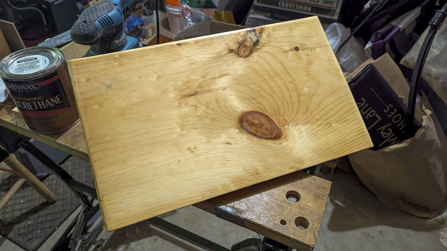 Rectangular wooden footstool being finished on a workshop bench beside a can of polyurethane and a power sander