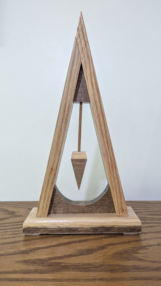 Triangular wooden sculpture with a pointed pendulum suspended above a curved base