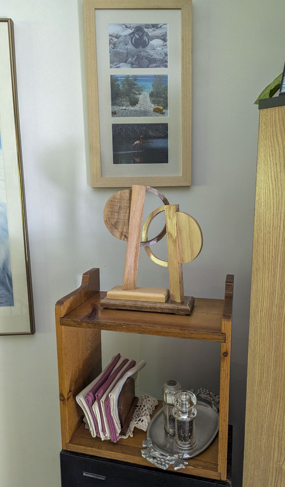 The abstract overlapping circle sculpture sitting on a wooden shelf underneath a frame with 3 pictures in it.