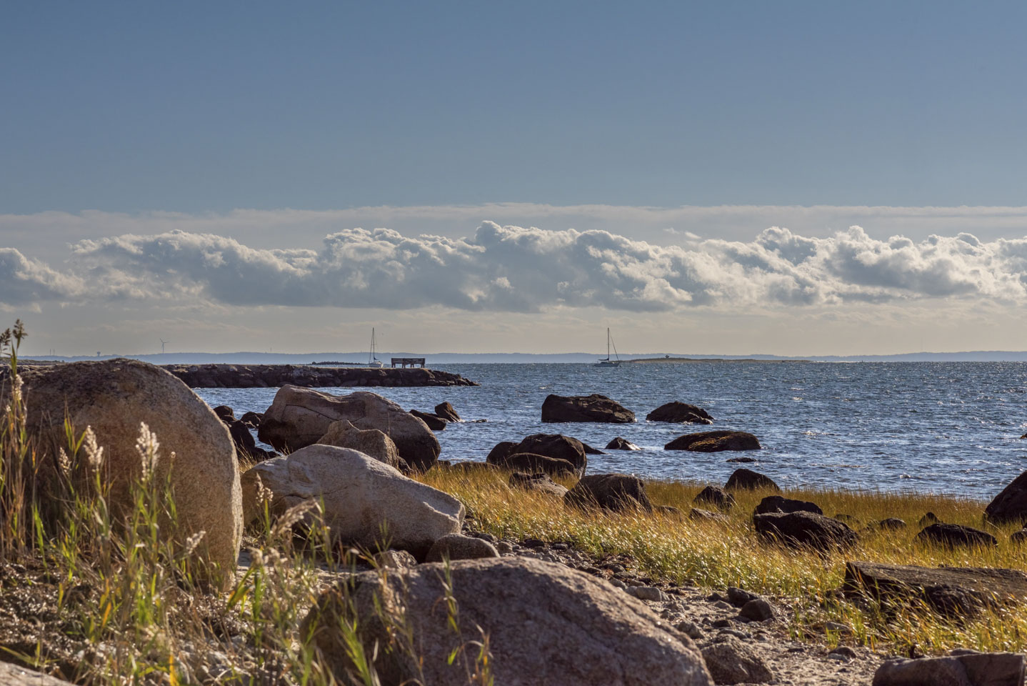 Rocky shore in the foreground with water and a couple of sailboats.