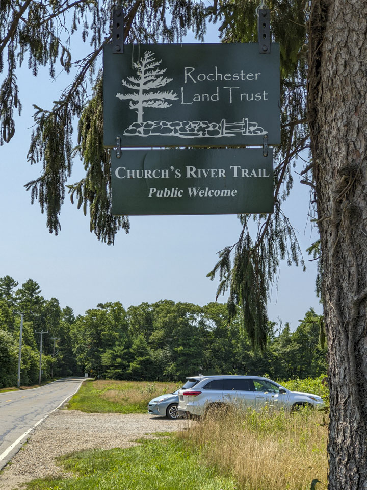 A sign for the preserve hangs from a tree, with a couple vehicles in the parking lot
